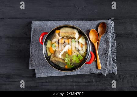 Soupe claire de brochet avec des légumes, du persil et des épices dans un pot rouge sur une table en bois noir avec des cuillères en bois, vue horizontale d'en haut, pose à plat Banque D'Images