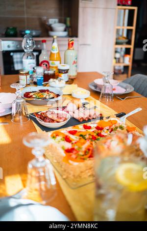 Un petit-déjeuner coloré sur une table de cuisine ensoleillée comprend des plats variés, des boissons, des viandes et des condiments, créant une ambiance chaleureuse et accueillante Banque D'Images