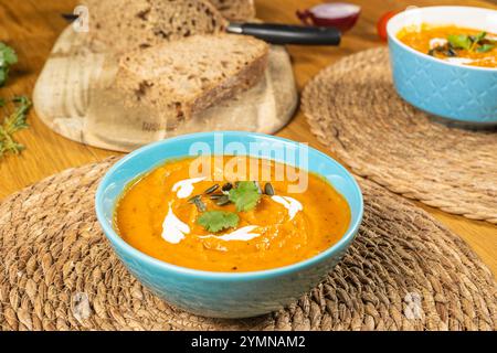 Des assiettes avec de la soupe de citrouille et des croûtons sur des stands écologiques texturés sont placées sur une table en bois, où des légumes, une citrouille, une planche à découper avec sl Banque D'Images