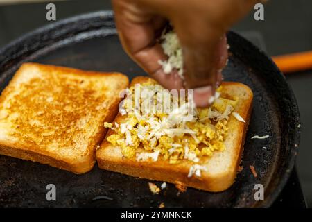 Mains ajoutant du fromage aux œufs brouillés sur du pain grillé dans une poêle. Petit déjeuner ou collation rapide et facile. Banque D'Images