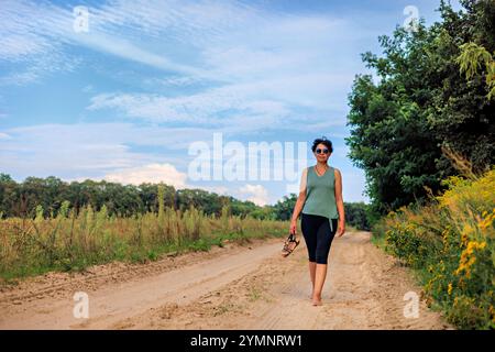 Une femme d'âge moyen marche pieds nus le long d'une route sablonneuse. Il y a un champ et des arbres à proximité. Banque D'Images