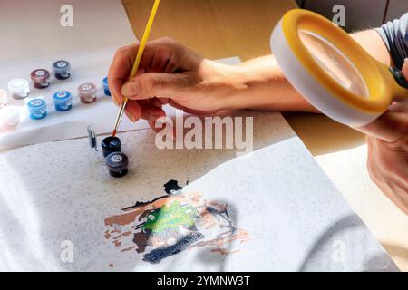 Peinture par chiffres. Une femme peint une peinture par chiffres avec un pinceau, regardant la toile à travers une loupe. Peintures dans des récipients en plastique Banque D'Images
