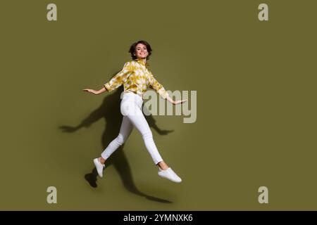 Jeune femme sautant joyeusement sur fond kaki, portant chemisier élégant et pantalon blanc, cheveux bruns volant, capturé à mi-mouvement. Banque D'Images