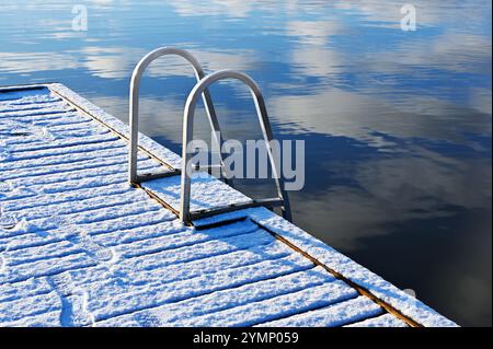 une vue rapprochée d'un quai en bois recouvert de neige avec une échelle métallique menant à l'eau calme et réfléchissante. La neige immaculée révèle de faibles empreintes de pas, ajoute Banque D'Images