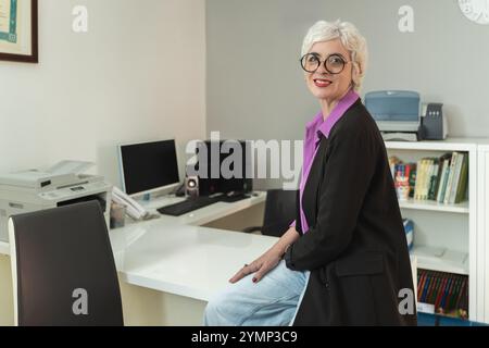 Femme d'affaires mature confiante avec coupe de cheveux pixie blanc et lunettes souriant tout en se tenant debout au bureau dans l'espace de travail de bureau moderne Banque D'Images