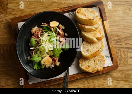 Un gros plan d'un plat gastronomique de tartare de bœuf servi dans un bol noir avec des herbes fraîches, garni d'œufs de caille et d'oignons, accompagné de plusieurs tranches de tartare Banque D'Images