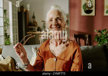 Femme âgée debout dans le salon, souriant tout en tenant des lunettes. Intérieur confortable avec des plantes, des œuvres d'art et des meubles modernes créant une atmosphère chaleureuse Banque D'Images