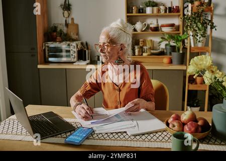 Femme senior assise au bureau examinant des documents avec ordinateur portable et smartphone entouré de plantes et de livres dans le bureau à domicile Banque D'Images