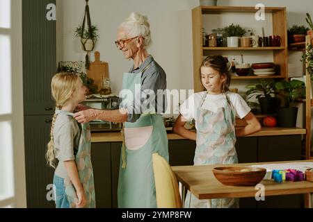 Femme âgée dans un tablier debout dans une cuisine moderne s'engageant avec deux enfants tout en préparant la nourriture. La cuisine est décorée avec des plantes et des éléments en bois créant une atmosphère chaleureuse Banque D'Images