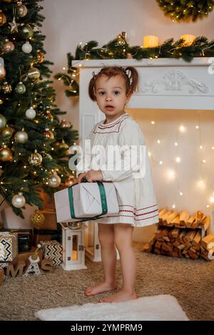 Petite fille assise près du sapin de Noël et ouvrant des cadeaux. Vacances d'hiver. Banque D'Images