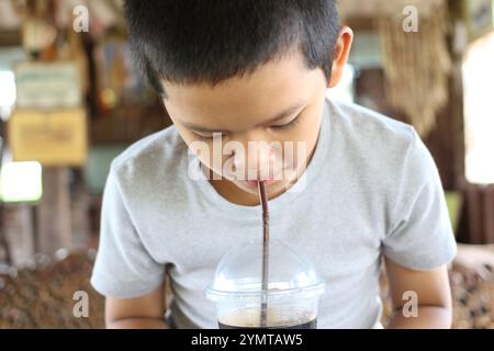 Un jeune garçon est assis à l'intérieur, sirotant une boisson rafraîchissante à travers une paille. L'atmosphère chaleureuse met en valeur la joie de l'enfance et les plaisirs simples de l'été. Banque D'Images