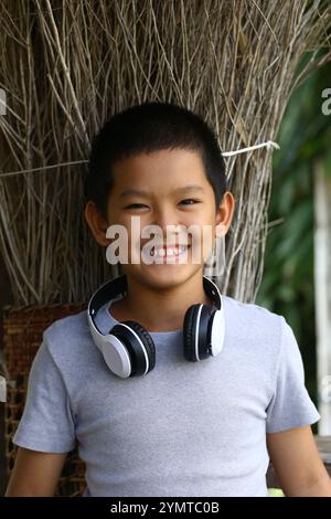 Un garçon joyeux portant des écouteurs sourit avec éclat sur un fond naturel, capturant l'essence de la jeunesse et du plaisir dans un cadre ludique. Banque D'Images