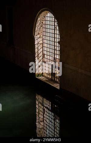 Vue panoramique sur le canal avec porte en fer la nuit à la vieille ville de Venise. Un faisceau lumineux jaune brille à travers la porte en fer. Eau verte. Banque D'Images