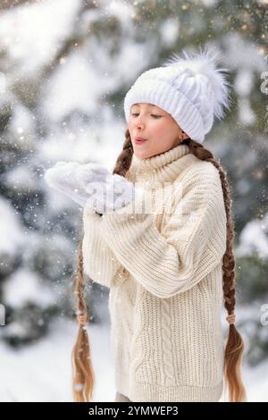 Une petite fille joyeuse dans un chapeau tricoté blanc se tient sur une route enneigée dans une forêt d'hiver calme de pin, et tient une grande boule de neige froide Banque D'Images