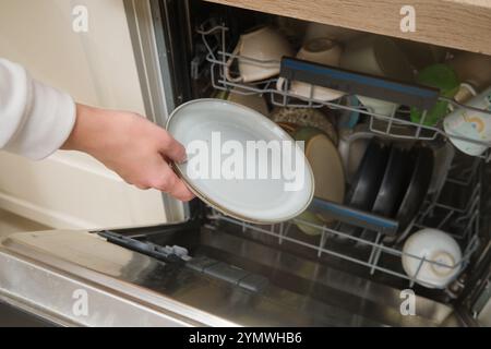 Main placer une assiette sale dans un lave-vaisselle rempli de vaisselle variée. Vue rapprochée du processus de lavage de la vaisselle dans la cuisine domestique. Banque D'Images