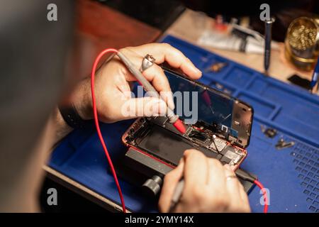 Technicien effectuant la réparation de la batterie d'un smartphone sur un appareil dans un atelier le soir Banque D'Images