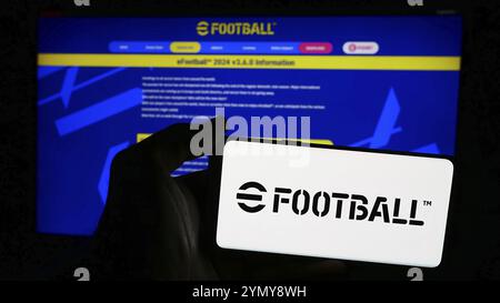 Stuttgart, Allemagne, 06-27-2024, personne tenant un téléphone portable avec le logo du jeu vidéo de football eFootball (Konami) devant la page web de l'entreprise. Focus o o Banque D'Images