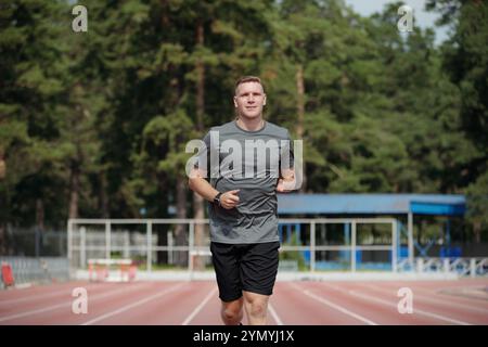 Homme qui fait du jogging sur une piste entourée d'arbres, portant une chemise grise et un short noir. Contexte comprend des installations sportives extérieures et une végétation luxuriante Banque D'Images
