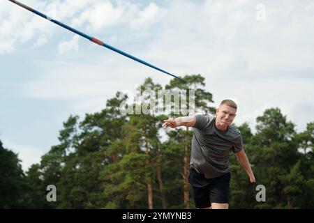 Athlète participant à une épreuve de lancer de javelot sur une piste extérieure. Arbres et ciel formant la toile de fond avec javelot dans les airs pendant le lancer Banque D'Images