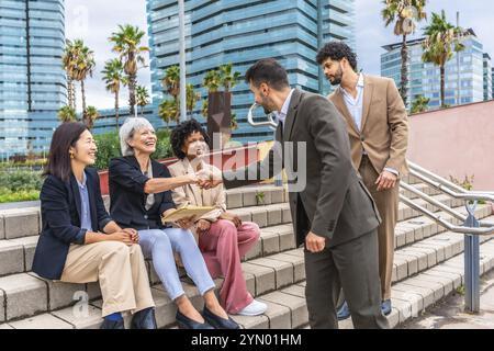 Homme d'affaires serrant la main avec une femme d'affaires mature assise à l'extérieur dans des escaliers urbains avec deux collègues féminins multi-ethniques Banque D'Images