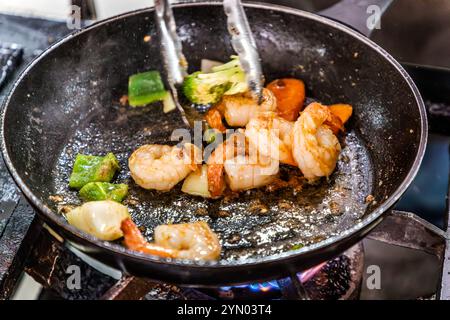 Les crevettes aux poivrons, brocoli et carottes grésillent dans une poêle sur une flamme de gaz ouverte – l’un des plats fraîchement préparés à Plasa Bieu, la halle du marché sur la De Ruyterkade à Willemstad, Curaçao. Les cuisines ouvertes offrent aux invités une vue directe sur la préparation des aliments. Plasa Bieu est un marché aéré et rustique avec des tables de pique-nique et des cuisines ouvertes servant des plats classiques de Curaçao. Banque D'Images