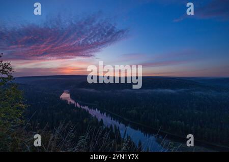 Alors que le soleil se couche à l'horizon, des teintes vibrantes remplissent le ciel au-dessus d'une rivière paisible serpentant à travers une forêt dense, créant une atmosphère sereine en soirée. Banque D'Images