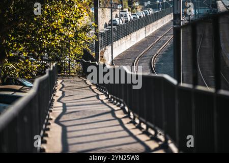 Une passerelle piétonne avec des garde-corps en métal projetant des ombres, à côté des voies de tramway et une route avec des voitures garées sous la lumière du soleil, entourée d'arbres à Sintr Banque D'Images