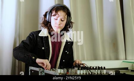 Une fille avec des écouteurs aime travailler sur un mixeur sonore, son expression concentrée et joyeuse. La photo met en lumière les thèmes de la production audio, de l'apprentissage Banque D'Images