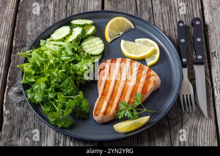 steak de thon grillé avec laitue fraîche, tranches de concombre et quartiers de citron sur une assiette gris foncé sur une table en bois rustique avec fourchette et couteau Banque D'Images