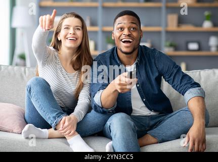 Couple multiracial joyeux s'amusant à la maison, regardant un film de comédie à la télévision Banque D'Images