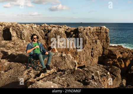 Homme se relaxant sur la plage rocheuse tout en profitant d'une boisson près de l'océan à la lumière du jour Banque D'Images