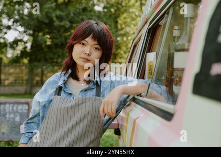 Gros plan moyen d'une belle ouvrière asiatique de camion de nourriture dans un tablier dépouillé s'appuyant sur une camionnette de couleur pastel Banque D'Images