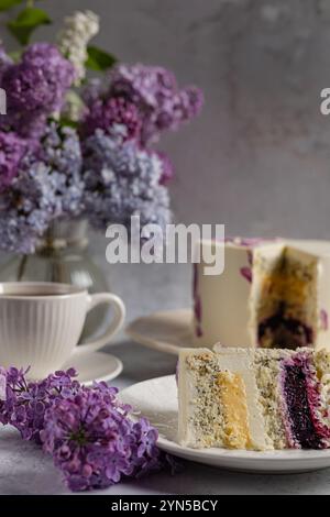Composition élégante avec une tranche de graines de pavot et un gâteau aux baies sur une assiette blanche, des fleurs de lilas et une tasse de thé, capturée dans une douce lumière naturelle Banque D'Images