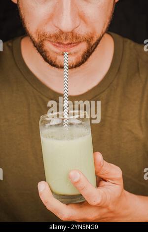 Gros plan d'un homme buvant du matcha latte vert dans un verre à travers une paille, concept de culture du thé. Un homme sirote du thé à travers une paille Banque D'Images