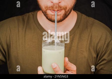 Gros plan d'un homme buvant du matcha latte vert dans un verre à travers une paille, concept de culture du thé. Un homme sirote du thé à travers une paille Banque D'Images