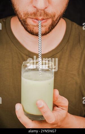 Gros plan d'un homme buvant du matcha latte vert dans un verre à travers une paille, concept de culture du thé. Un homme sirote du thé à travers une paille Banque D'Images