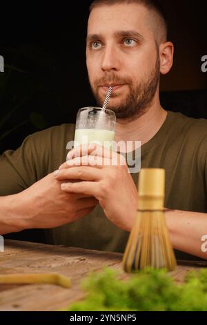 Un homme réfléchi apprécie un verre de matcha latte crémeux avec un fouet en bambou sur une table en bois. Idéal pour les thèmes de pleine conscience, de bien-être et de thé cultu Banque D'Images