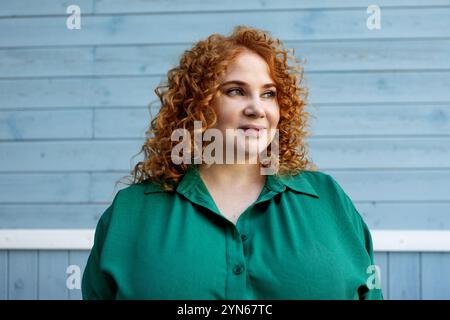Portrait plus taille Curvy femme avec cheveux rouges bouclés dans des vêtements verts contre fond de mur en bois bleu vibrant, beauté et confiance en extérieur Banque D'Images