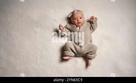 Un bébé européen actif, pleine longueur, âgé d'un mois, repose sur une couverture dans une combinaison tricotée avec un noeud sur la tête, de couleur pastel. Banque D'Images