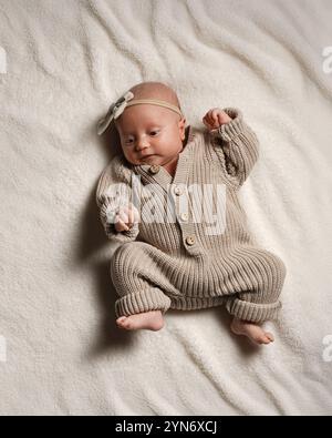 Un bébé européen actif, pleine longueur, âgé d'un mois, repose sur une couverture dans une combinaison tricotée avec un noeud sur la tête, de couleur pastel. Banque D'Images