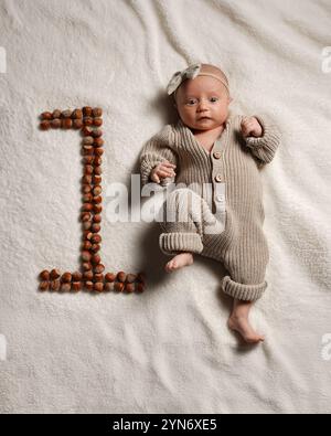 Bébé européen actif en pleine hauteur, âgé d'un mois, sur une couverture dans une salopette tricotée avec un arc sur la tête, le numéro est celui des noix naturelles, pastel Banque D'Images