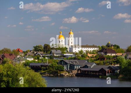 Une scène sereine au bord de la rivière avec une église au dôme doré, des maisons traditionnelles et une végétation luxuriante sous un ciel bleu vif avec des nuages éparpillés, se reflètent Banque D'Images