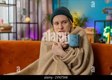 Une jeune femme couverte d'une couverture chaude boit du café à la maison. La femelle caucasienne gèle à cause du froid, se sent mal, malsaine. fille malade avec des symptômes de la grippe couverte de couverture se réchauffe avec du thé chaud à la maison. Banque D'Images