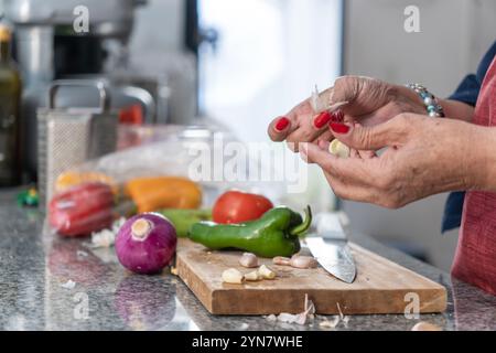 Une femme coupe des légumes sur une planche à découper. Elle porte du rouge et a des ongles rouges. Les légumes comprennent des poivrons, des oignons et de l'ail Banque D'Images