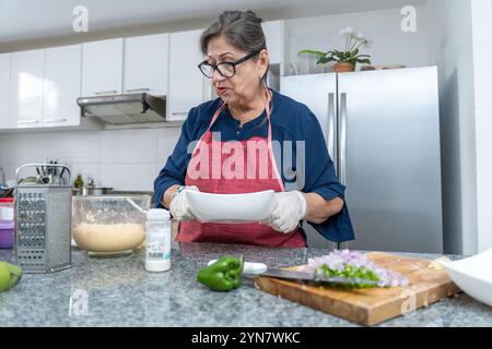 Une femme dans un tablier bleu prépare de la nourriture dans une cuisine. Elle porte des gants et tient une assiette blanche Banque D'Images