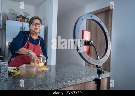 Femme Latina senior enregistrant un cours de cuisine sur vidéo avec une lumière et un téléphone portable pour les médias sociaux. Femme adulte moderne produisant du contenu numérique Banque D'Images