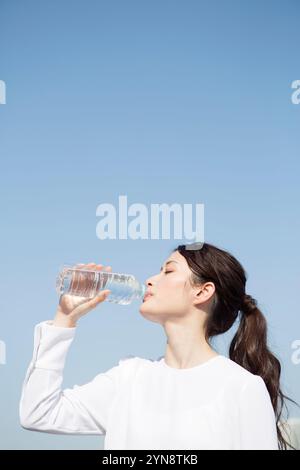 Femme dans la vingtaine buvant une bouteille PET sous un ciel bleu de profil Banque D'Images