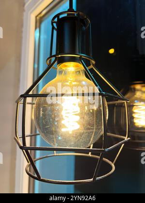 Une ampoule lumineuse incandescente dans un luminaire en cage métallique, mettant en valeur un design industriel moderne. L'éclairage chaleureux et ambiant crée une atmosphère chaleureuse, parfaite pour Banque D'Images