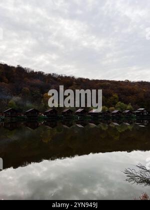 Une scène paisible au bord du lac avec des cabanes rustiques reflétant dans l'eau calme, entouré par une forêt d'automne animée sous un ciel nuageux. Banque D'Images