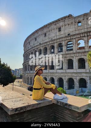 Vêtu d'une tenue jaune éclatante, un voyageur est assis sur un mur de pierre, admirant le majestueux Colisée de Rome sous un ciel bleu clair. La lumière chaude du soleil améliore la beauté de la structure ancienne. Banque D'Images